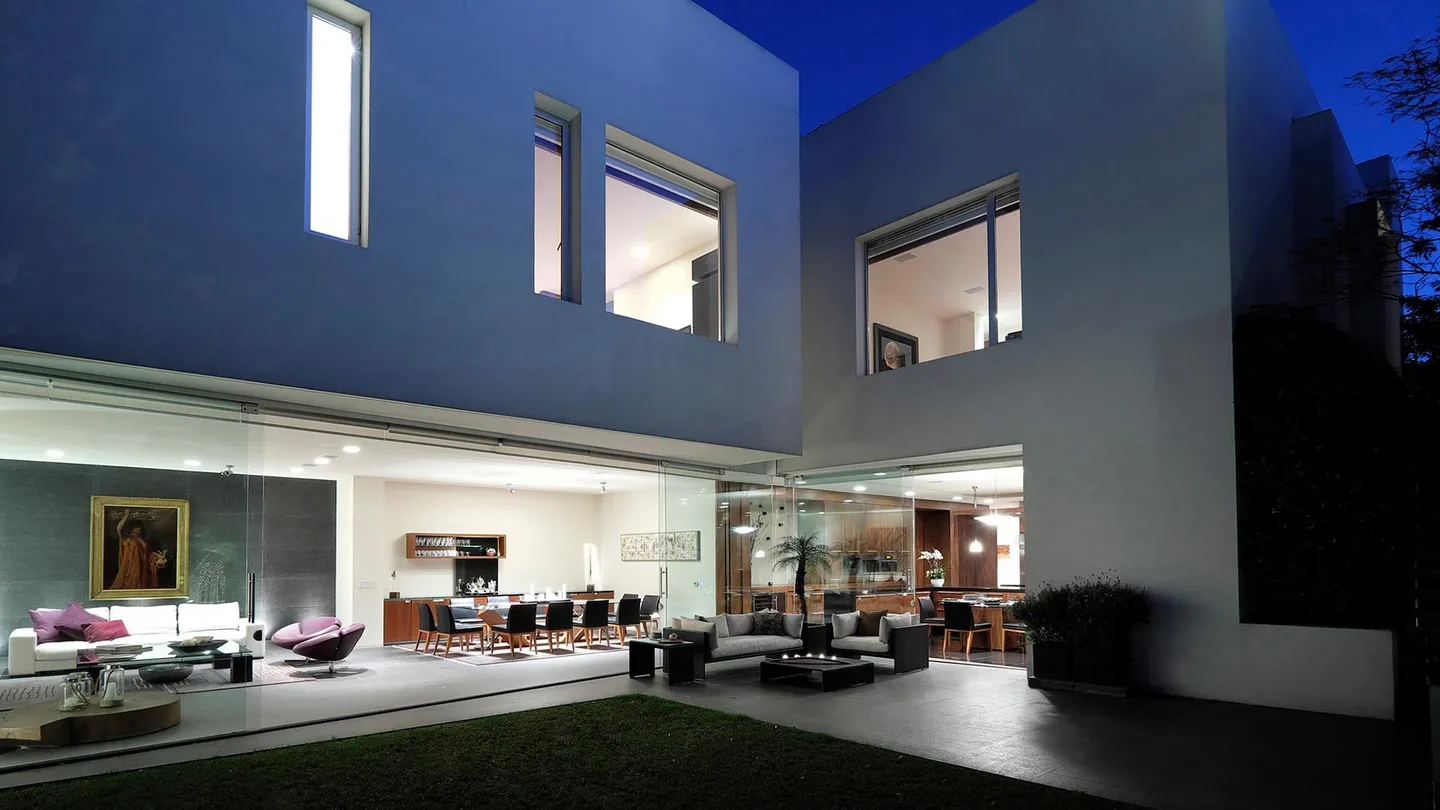 Casa FU3: estancia abierta a la terraza bajo volúmenes de muros oscuros, en la hora azul