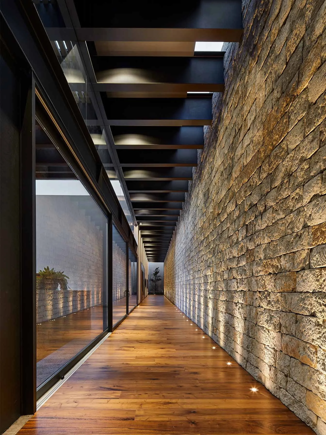 Casa IG: pasillo de madera entre un muro de piedra y un ventanal