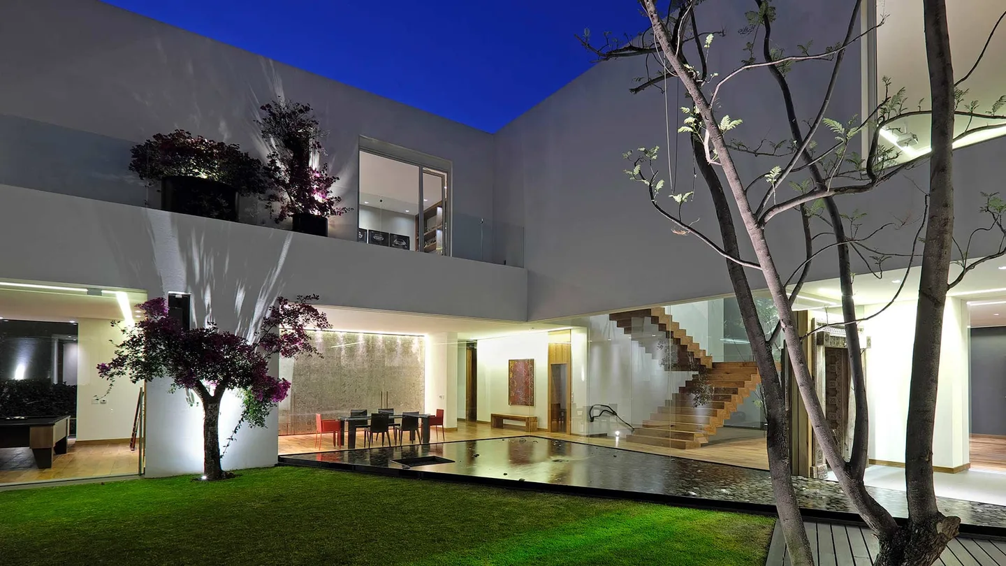 Casa LV: patio ajardinado al anochecer con la escalera de madera iluminada tras el vidrio