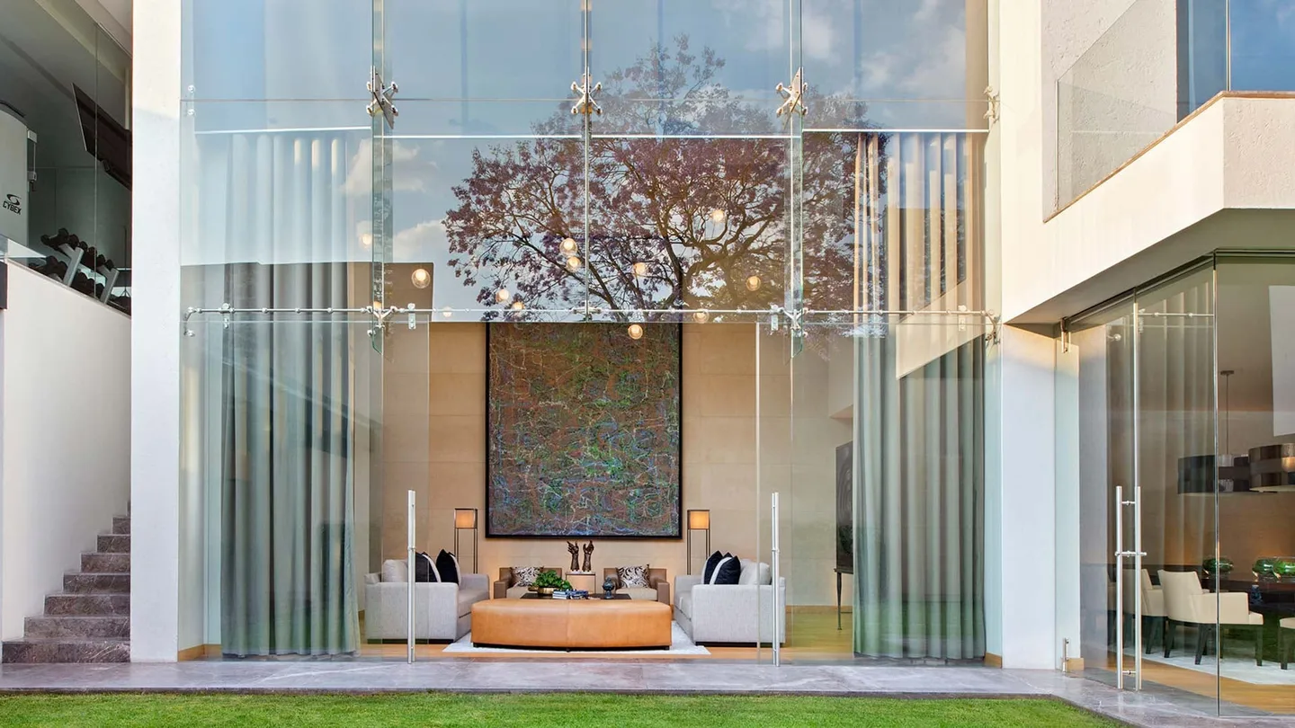 Casa OC: estancia a doble altura tras un muro de vidrio, con una jacaranda reflejada