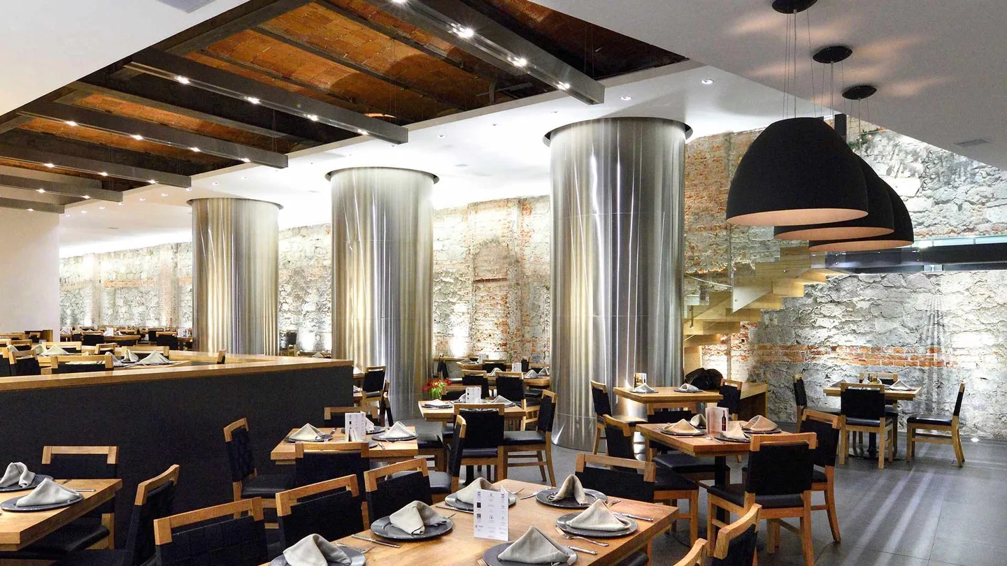 Restaurante 5M: columnas metálicas y lámpara suspendida frente al muro de piedra