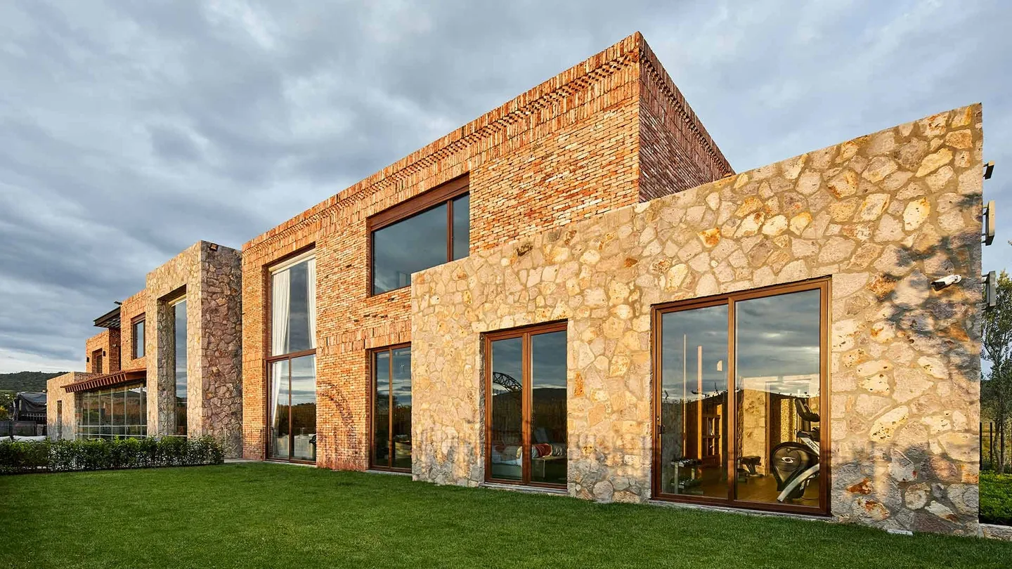 Casa SM: volúmenes de piedra y ladrillo sobre el jardín bajo un cielo nublado