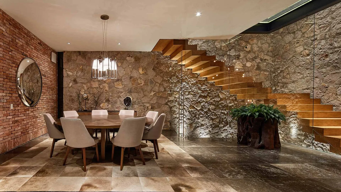 Casa SM: comedor con escalera de madera junto a un muro de piedra