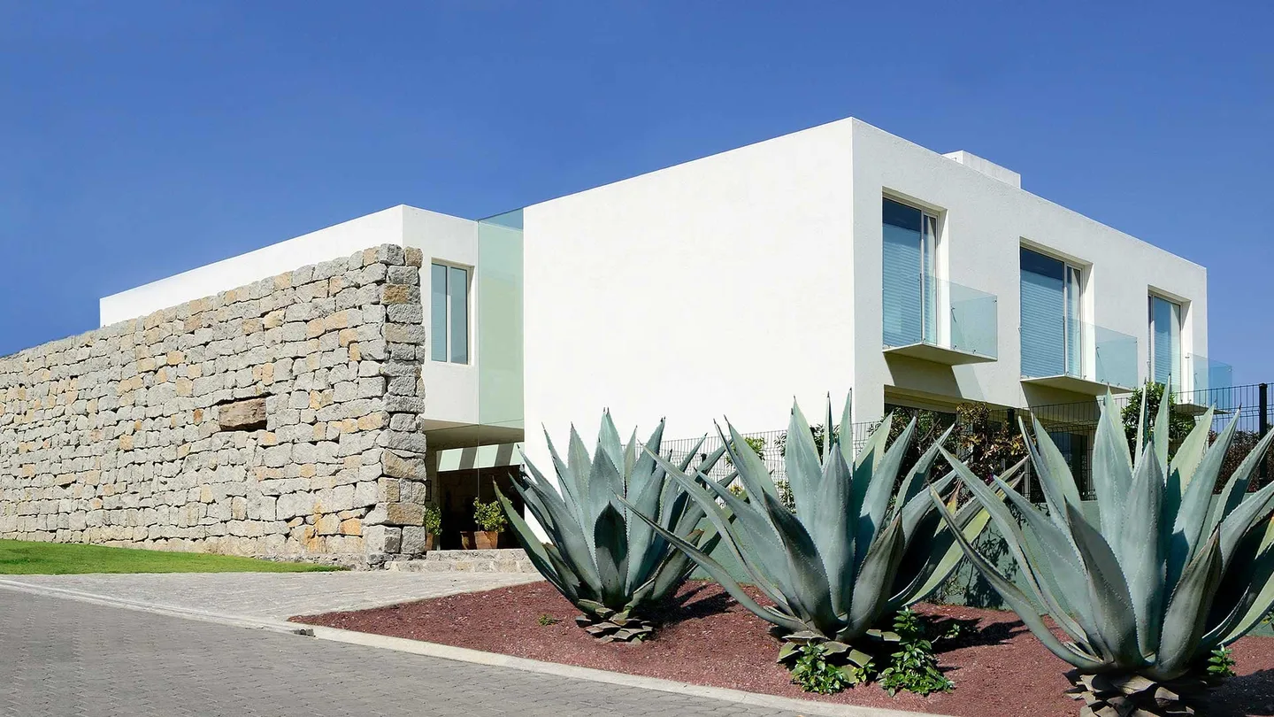 Casa VE: muro de piedra y volumen blanco con agaves en primer plano