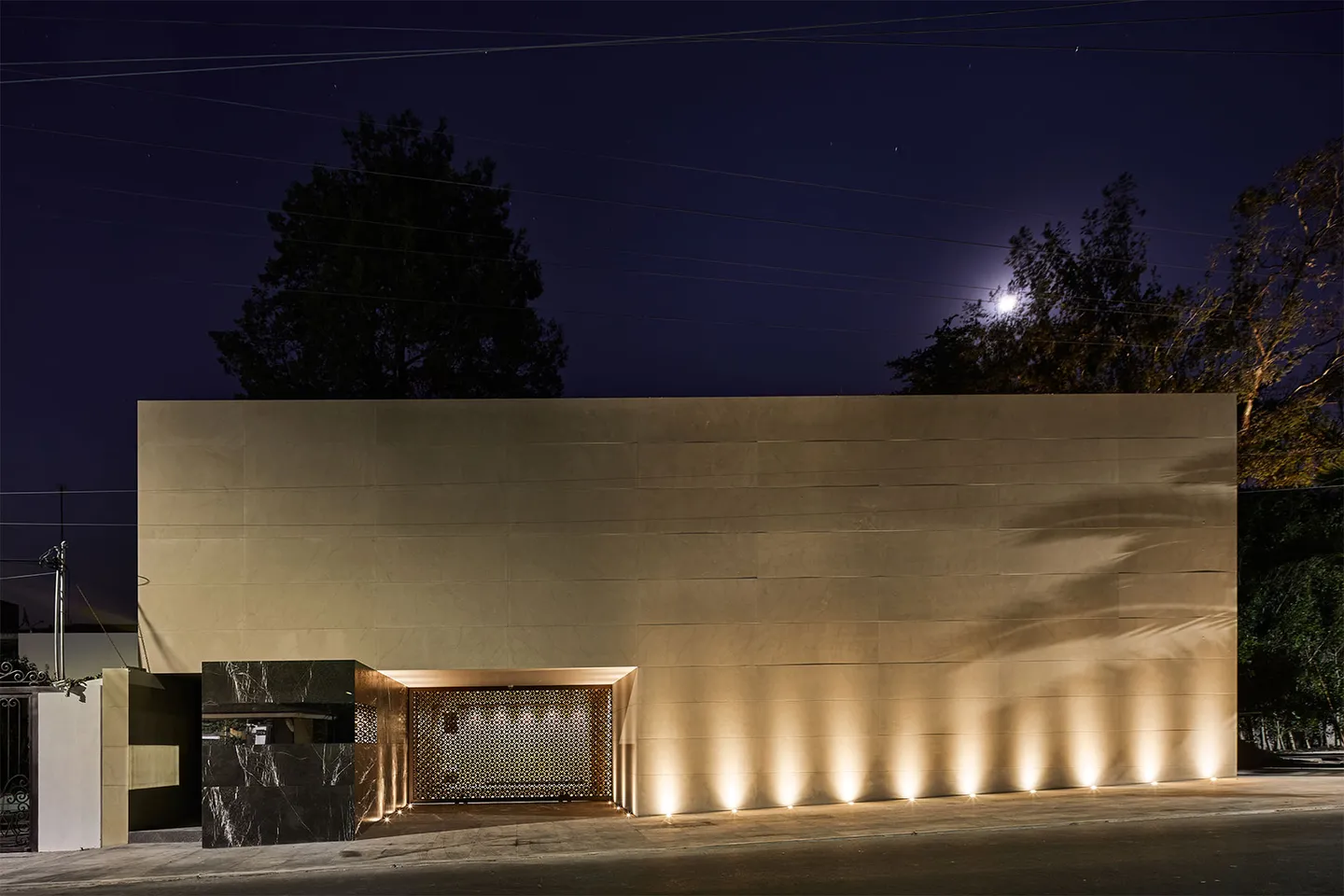 Casa de noche: el muro de piedra iluminado desde el suelo, con la luna sobre los árboles